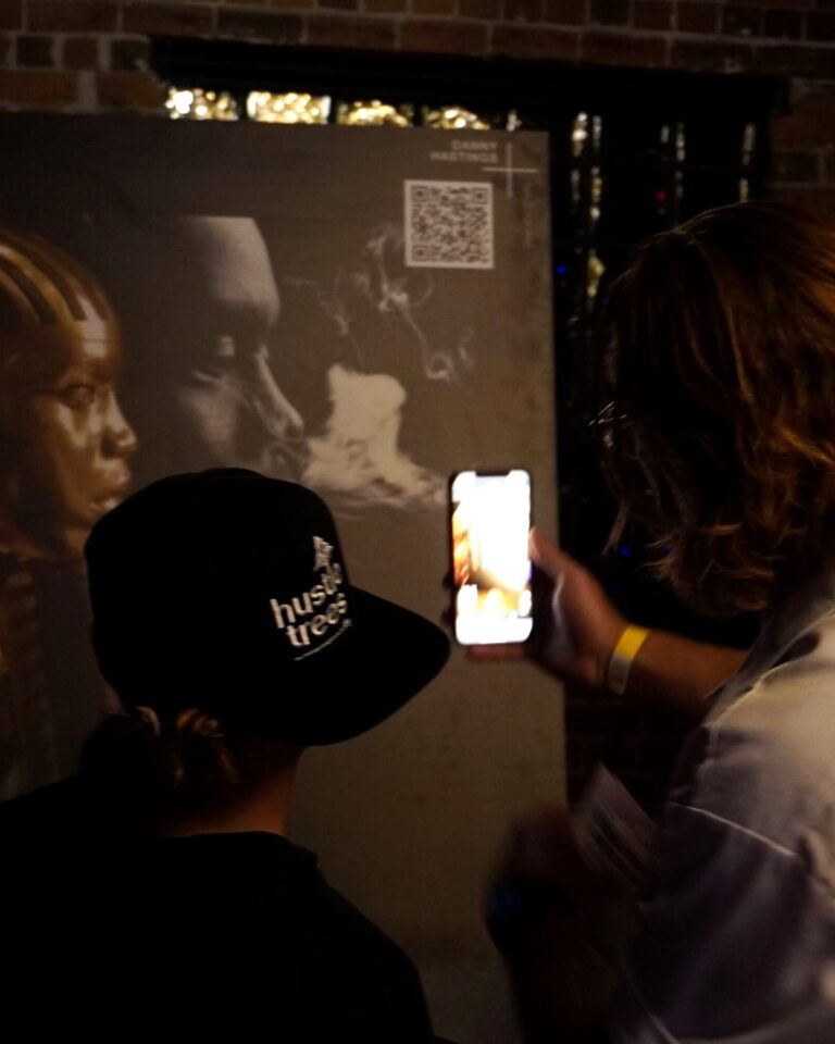Two visitors at Cultur-ill with Curaleaf in Phoenix scan the QR code printed in the corner of an artwork, and the AR opens on the phone