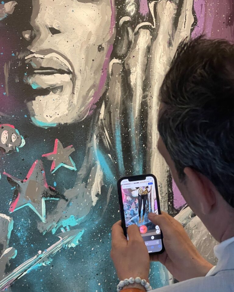 A visitor at Zany Studios Art Gallery holds up a phone in front of a painting after scanning its QR code, with the AR showing on screen