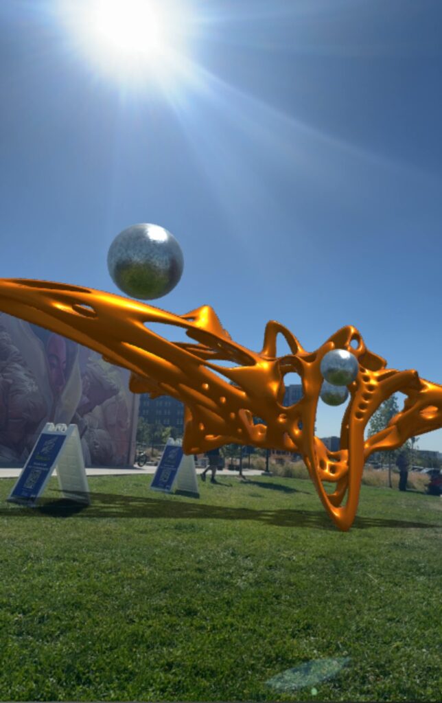 An orange, flowing AR sculpture with a chrome sphere hovering over the grass in a park at Denver Walls 2024