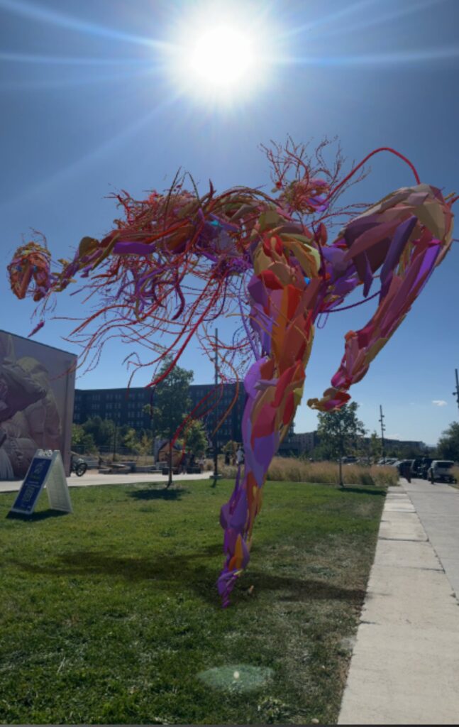 A red, sinewy AR sculpture standing beside a park path in the sun at Denver Walls 2024